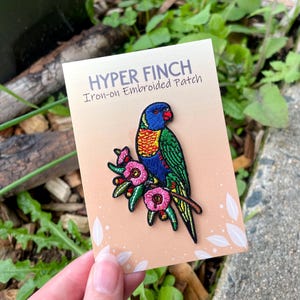May include: An iron-on embroidered patch with a vibrant parrot design. The patch shows a parrot with blue, green, yellow, and red plumage, perched on a branch with pink flowers. The text "HYPER FINCH" and "Iron-on Embroidered Patch" are displayed.