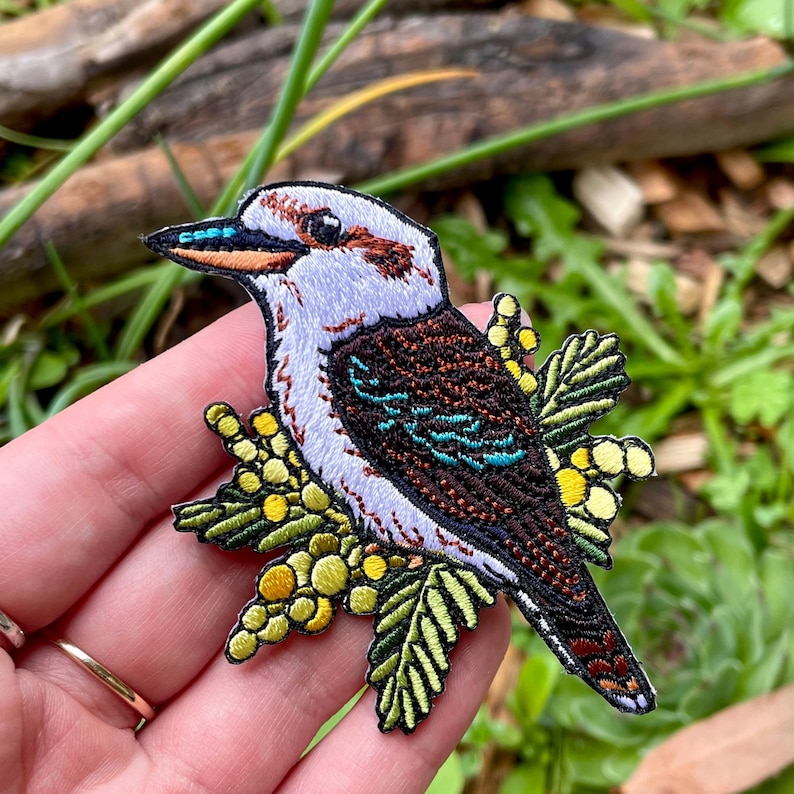 May include: Embroidered patch of a kookaburra bird with a white head, brown wings, and teal details. The bird is set on yellow berries and green leaves. The patch is designed to be sewn or ironed onto fabric.