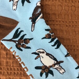 May include: A pair of blue socks with a white kookaburra bird and green gum leaves pattern. The socks have the text "Kookaburra" printed on them.