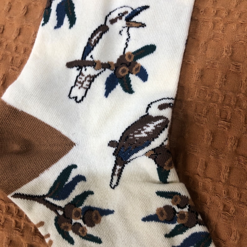 May include: White socks with a brown heel and toe. The socks feature a repeating pattern of kookaburras, native Australian birds, perched on branches with gum nuts.