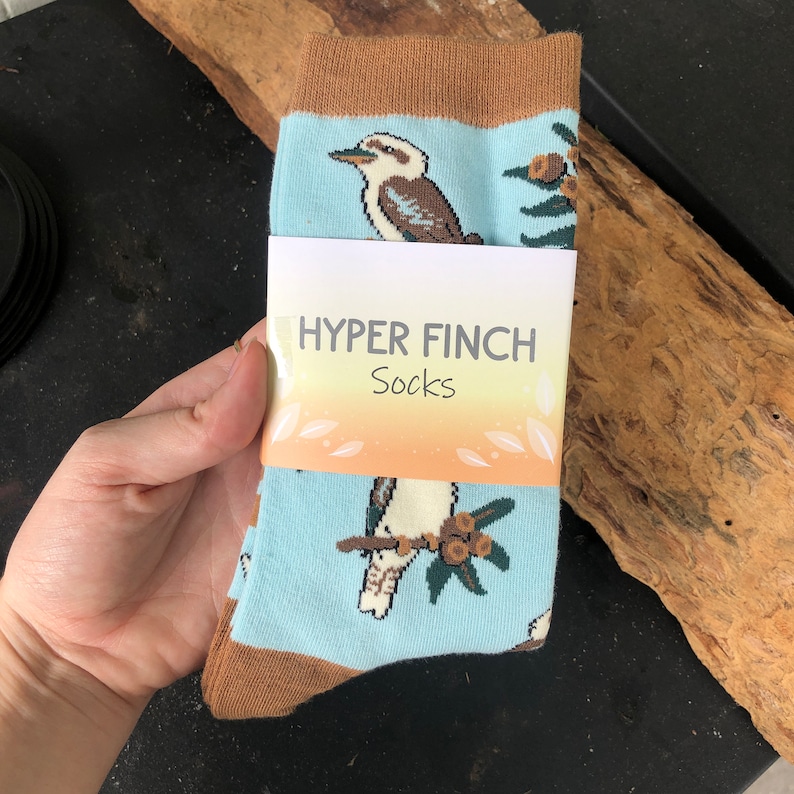 May include: A pair of blue socks with a brown cuff. The socks feature a white bird with brown markings and a green leafy branch with brown berries. The socks have a white tag that reads "HYPER FINCH Socks".