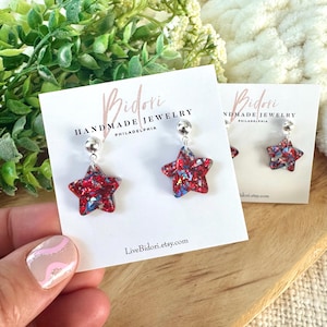 May include: Star-shaped earrings with red, white, and blue glitter embedded in clear resin. The earrings are attached to silver-colored posts. The earrings are displayed on a white card with the text "Bidori Handmade Jewelry Philadelphia" and "LiveBidori.etsy.com".