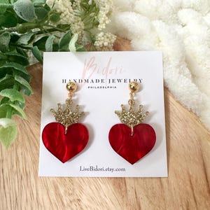 Pode incluir: Brincos em forma de cora&ccedil;&atilde;o vermelho com coroas douradas com glitter. Os brincos est&atilde;o em um cart&atilde;o branco com o texto "Bidori Handmade Jewelry Philadelphia" e "LiveBidori.etsy.com".