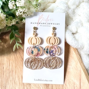 May include: Pumpkin-shaped dangle earrings. The earrings feature a layered design with cream-colored pumpkin tops, floral-patterned centers, and gold glitter pumpkin bottoms. The earrings are displayed on a white card with the text "Bidori Handmade Jewelry Philadelphia" and the Etsy shop URL.