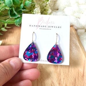 Puede incluir: Pendientes en forma de lágrima con ganchos dorados, rellenos de purpurina morada, azul y rosa. Los pendientes se exhiben en una tarjeta blanca con el texto "Bidori Handmade Jewelry Philadelphia".