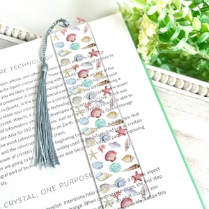 May include: A bookmark with a watercolor illustration of seashells and starfish on a white background. The bookmark has a blue tassel and text that reads "E Crystal, One Purpose" and "Practice = Transformation".