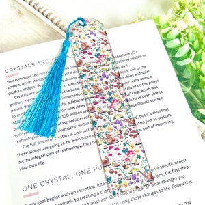 May include: A bookmark with a colorful design featuring snails, mushrooms, and flowers. The bookmark is made of glass and has a blue tassel. The text on the bookmark reads "One Crystal, One Purpose".