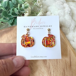 Puede incluir: Un par de pendientes con forma de calabaza con postes dorados. Los pendientes son de color naranja y amarillo con detalles de purpurina. Se muestran en una tarjeta blanca con el texto "Bidori Handmade Jewelry Philadelphia" y el sitio web LiveBidori.etsy.com.