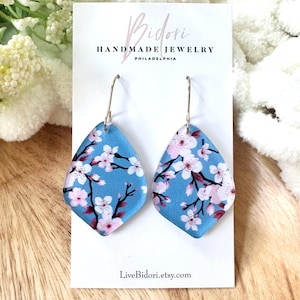 May include: A pair of blue, diamond-shaped earrings with a floral cherry blossom design. The earrings have gold-colored hooks and are displayed on a white card with the text "Bidori Handmade Jewelry Philadelphia" and "LiveBidori.etsy.com".