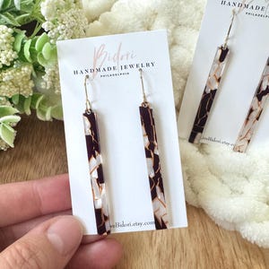 May include: Rectangular earrings with a marbled design in shades of burgundy, white, and gold. The earrings are displayed on a white card with the text "Bidori Handmade Jewelry Philadelphia". The earrings have gold-colored hooks.