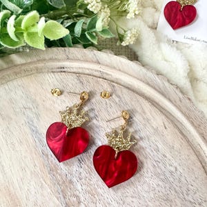 Pode incluir: Um par de brincos de cora&ccedil;&atilde;o vermelho com coroas douradas de glitter. Os brincos est&atilde;o sobre uma superf&iacute;cie de madeira.