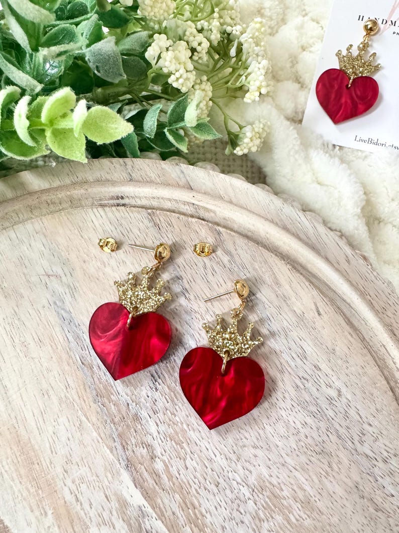 Pode incluir: Um par de brincos de cora&ccedil;&atilde;o vermelho com coroas douradas de glitter. Os brincos est&atilde;o sobre uma superf&iacute;cie de madeira.