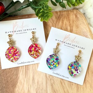May include: Two pairs of handmade earrings on white cards. Each earring features a gold star stud and a clear, teardrop-shaped pendant filled with colorful glitter. The cards display the brand name "Bidori Handmade Jewelry" and the website address.