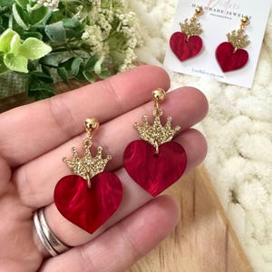 Pode incluir: Brincos em forma de cora&ccedil;&atilde;o vermelho com coroas douradas de glitter. Os brincos s&atilde;o feitos de acr&iacute;lico e t&ecirc;m um efeito marmorizado.