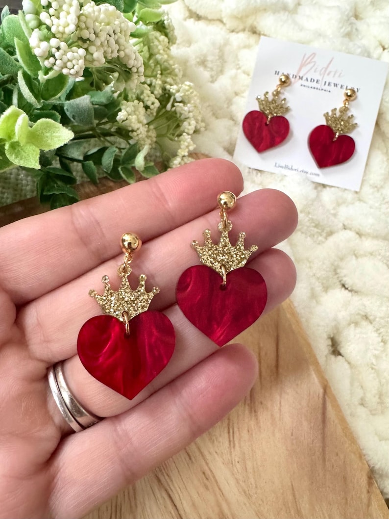 Pode incluir: Brincos em forma de cora&ccedil;&atilde;o vermelho com coroas douradas de glitter. Os brincos s&atilde;o feitos de acr&iacute;lico e t&ecirc;m um efeito marmorizado.