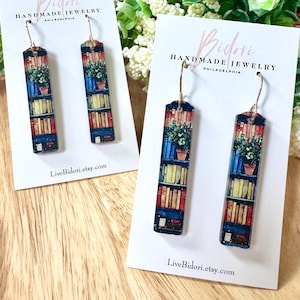 May include: A pair of rectangular earrings featuring a bookshelf design with books, a plant, and a starry night sky. The earrings are displayed on white cards with the text "Bidori Handmade Jewelry Philadelphia" and a website address.