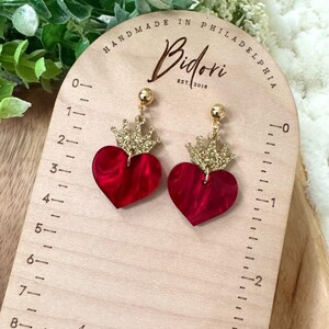 Pode incluir: Um par de brincos em forma de cora&ccedil;&atilde;o vermelho com coroas douradas de glitter. Os brincos est&atilde;o em uma r&eacute;gua de madeira com o texto "HANDMADE IN PHILADELPHIA Bidori EST 2018" impresso nela.