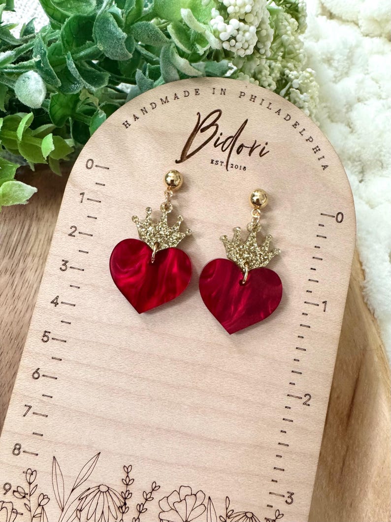 Pode incluir: Um par de brincos em forma de cora&ccedil;&atilde;o vermelho com coroas douradas de glitter. Os brincos est&atilde;o em uma r&eacute;gua de madeira com o texto "HANDMADE IN PHILADELPHIA Bidori EST 2018" impresso nela.