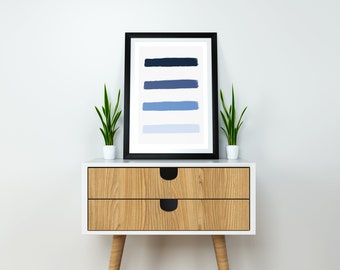 May include: A framed abstract print with five horizontal blue brushstrokes of varying shades, from dark blue to light blue, on a white background. The print is displayed on a white nightstand with two drawers and wooden legs, with a green plant on each side.