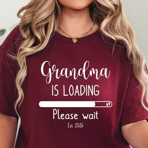 May include: Burgundy t-shirt with the text "Grandma is Loading" in white script. Below the text is a loading bar graphic and the words "Please wait" and "Est. 2026". The shirt is a short-sleeved crew neck.