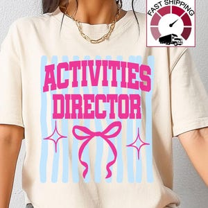 May include: A cream-colored short-sleeve t-shirt featuring the text "ACTIVITIES DIRECTOR" in bold pink letters. The design includes light blue vertical stripes, a pink bow graphic, and two four-pointed star icons centered on the chest area.