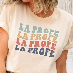 May include: A light beige t-shirt with the text "LA PROFE" repeated four times in a rainbow color scheme. The text is in a retro font style.
