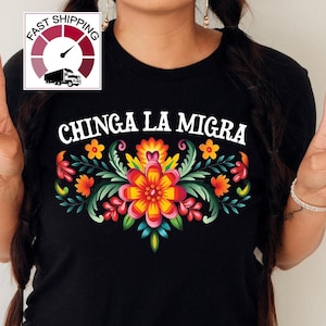 May include: Black t-shirt with the text "CHINGA LA MIGRA" above a colorful floral design. The design features orange, yellow, red, and green flowers and leaves. The shirt has a round neck.