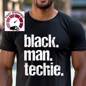 May include: Black crew neck t-shirt with the text "black. man. techie." in a white, distressed font. The shirt is worn by a person with a blurred background.