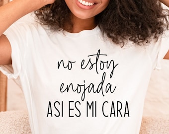 May include: A white t-shirt with black text that reads "no estoy enojada asi es mi cara". The person wearing the shirt is smiling and has curly hair.