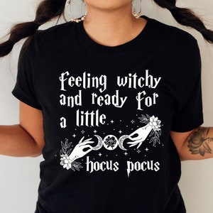 May include: Black t-shirt with white text that reads "Feeling witchy and ready for a little hocus pocus". The text is surrounded by a floral design and a crescent moon.