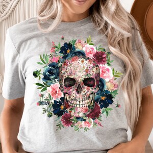 May include: A light gray t-shirt featuring a floral skull design. The skull is adorned with pink, blue, and white flowers, surrounded by a wreath of dark blue and burgundy blooms. The shirt is a crew neck style.
