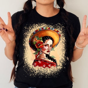 May include: Black t-shirt with a white splatter design featuring a woman with dark hair wearing a large, yellow sombrero with red and yellow flowers.