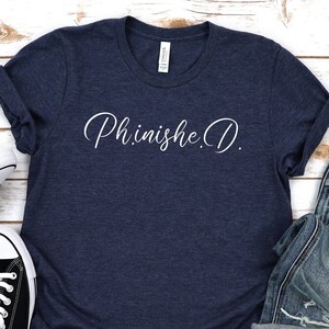 May include: A navy blue t-shirt with the text "Phinishe.D." in white script on the front. The shirt is laid flat on a white wooden surface with a pair of black sneakers and a pair of blue jeans.