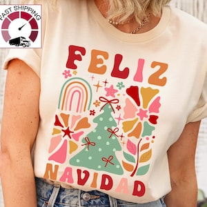 May include: A cream-colored t-shirt with a colorful graphic design featuring the text "Feliz Navidad" in a retro style. The design includes a Christmas tree, flowers, and a rainbow.