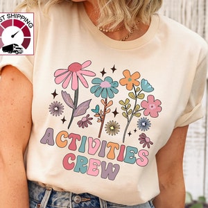 May include: A beige t-shirt with a colorful floral design and the text "ACTIVITIES CREW" in a retro font.