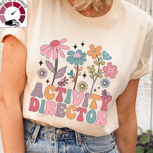May include: A cream colored t-shirt with a colorful graphic of flowers and the text "ACTIVITY DIRECTOR" in a retro font.