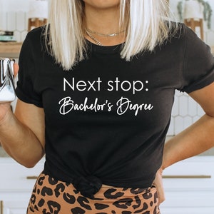 May include: A black t-shirt with the text "Next stop: Bachelor's Degree" in white lettering. The shirt is worn by a person holding a black and white marbled mug.