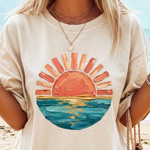 May include: A beige short-sleeved t-shirt featuring a circular graphic of a sun setting over the ocean. The design uses thick, textured paint strokes in shades of orange, yellow, and blue to depict the sun, sky, and water in a painterly style.