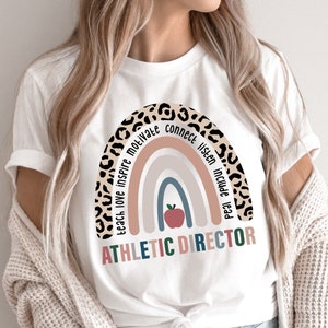 May include: White t-shirt with a rainbow design and the words "Teach Love Inspire Motivate Connect Listen Include Lead Athletic Director". The rainbow is made of different colors and has a leopard print border. There is a red apple in the center of the rainbow.