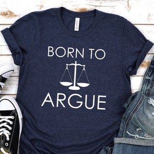 May include: A navy blue t-shirt with white text that reads "Born to Argue" and a white scale graphic. The t-shirt is laid out on a white wooden surface with a pair of black sneakers and a pair of blue jeans.
