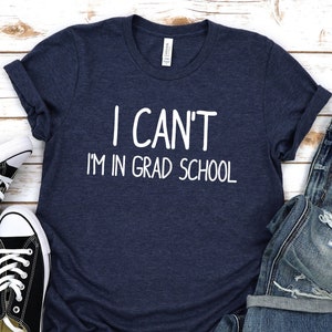 May include: A navy blue t-shirt with the text "I CAN'T I'M IN GRAD SCHOOL" in white. The shirt is laid out on a white wooden surface with a pair of black sneakers and blue jeans.