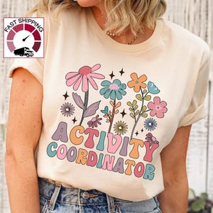 May include: A cream-colored t-shirt featuring a colorful floral design and the words "ACTIVITY COORDINATOR" in a retro font. The design includes pink, blue, orange, and green flowers. The shirt is made of a soft material.