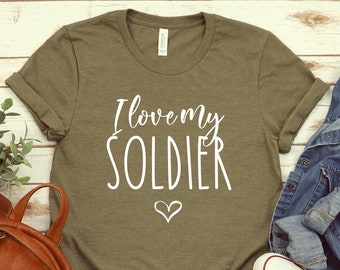 Puede incluir: Una camiseta verde con el texto "I love my SOLDIER" en letras blancas y un pequeño corazón blanco. La camiseta está sobre una superficie de madera blanca con una mochila de cuero marrón, un par de zapatillas blancas y una chaqueta vaquera azul.