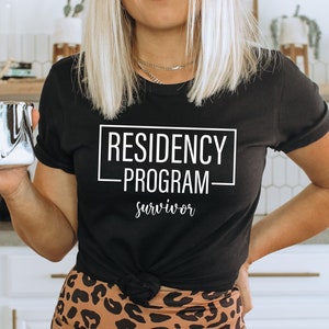 May include: Black t-shirt with the text "RESIDENCY PROGRAM survivor" in a white rectangular box. The shirt is worn with leopard print pants and a silver mug with a black and white swirl design.
