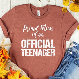 May include: A rust-colored t-shirt with the text "Proud Mom of an Official Teenager" in white. The shirt is laid out on a white wooden surface with a pair of blue jeans and fall-themed decorations.