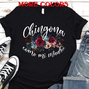 Puede incluir: Camiseta negra con texto blanco que dice "Chingona como mi madre" con un dise&ntilde;o floral. La camiseta est&aacute; colocada sobre una superficie de madera blanca con un par de zapatillas negras y unos vaqueros azules.