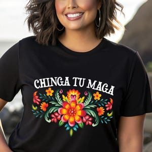 May include: Black t-shirt with the text "CHINGA TU MAGA" above a colorful floral design. The design features red, orange, yellow, and green flowers. The shirt is short-sleeved.
