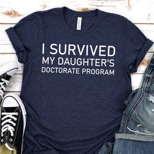 May include: A navy blue t-shirt with white text that reads "I survived my daughter's doctorate program". The shirt is laid out on a white wooden surface with a pair of black sneakers and a pair of blue jeans.