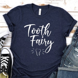 May include: A navy blue t-shirt with the text "Tooth Fairy" in white script lettering. A white tooth graphic is below the text. The shirt is laid out on a white wooden surface with a pair of black sneakers and a pair of blue jeans.
