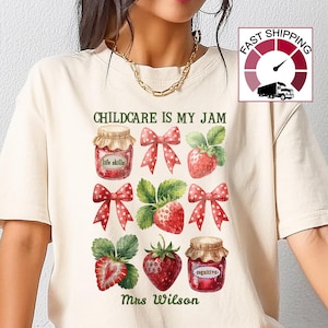 May include: Cream-colored t-shirt featuring a graphic design with the text "CHILDCARE IS MY JAM" and illustrations of strawberry jam jars, strawberries, and red bows. The name "Mrs. Wilson" is printed below the design.
